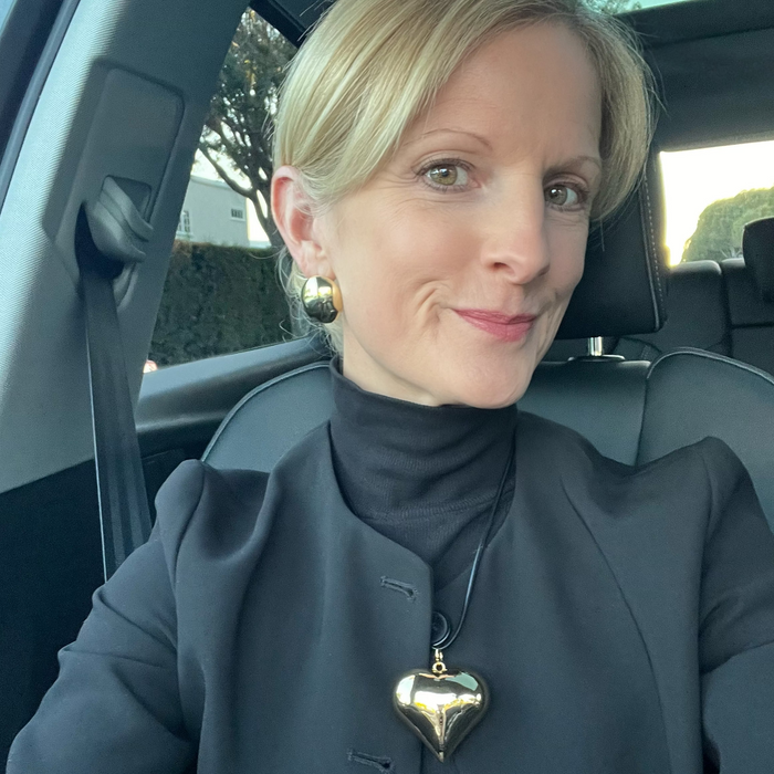 Woman sitting in a car wearing a dark coat with a heart-shaped pendant.