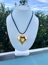 Gold heart-shaped pendant on a blue cord displayed on a white mannequin bust with a blurred outdoor background.