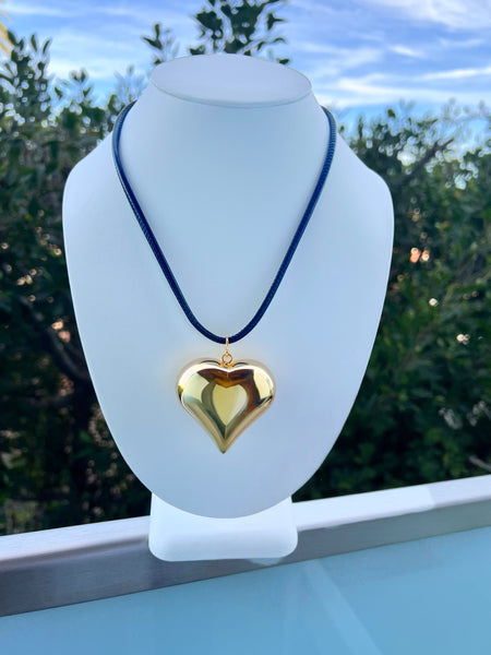 Gold heart-shaped pendant on a blue cord displayed on a white mannequin bust with a blurred outdoor background.