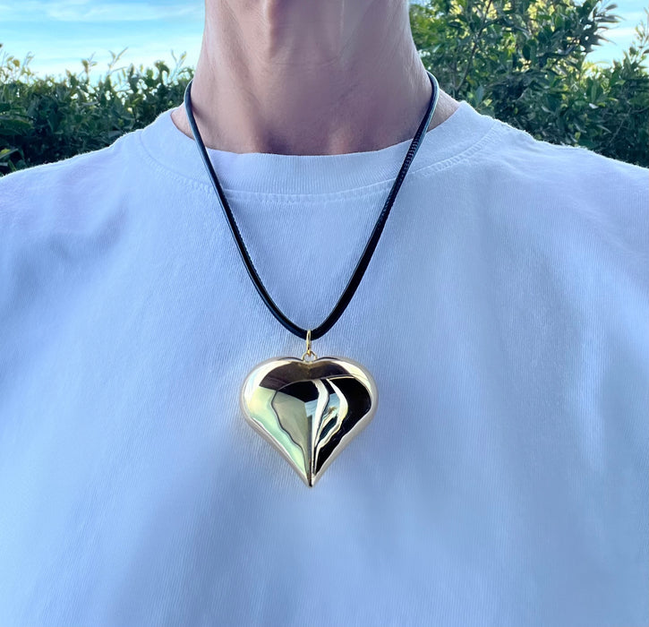 Person wearing a gold heart-shaped pendant on a black cord against a natural background