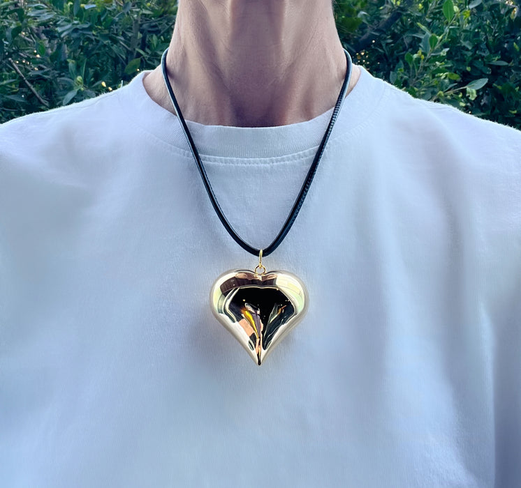 Person wearing a gold heart-shaped pendant on a black cord against a blurred green outdoor background