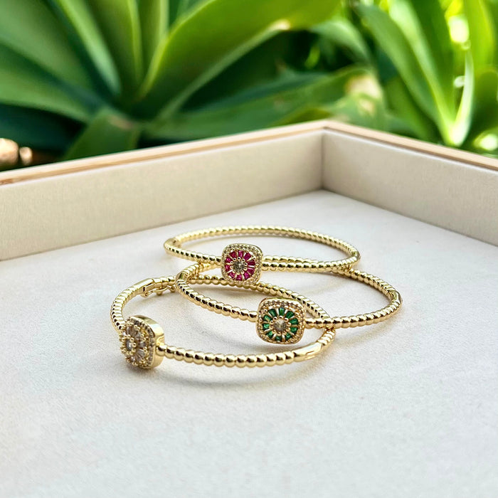 Gold bracelets with gemstones on a white surface with green foliage in the background