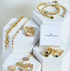 Gold jewelry set including necklaces, bracelets, and rings on white hexagonal stands with 'CF' branding.
