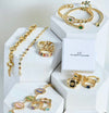 Gold jewelry set including necklaces, bracelets, and rings on white hexagonal stands with 'CF' branding.