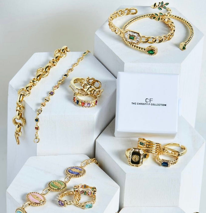 Gold jewelry set including necklaces, bracelets, and rings on white hexagonal stands with 'CF' branding.