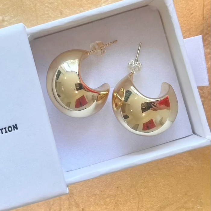 A pair of gold crescent-shaped hoop earrings displayed inside a white jewelry box on a light wood surface.