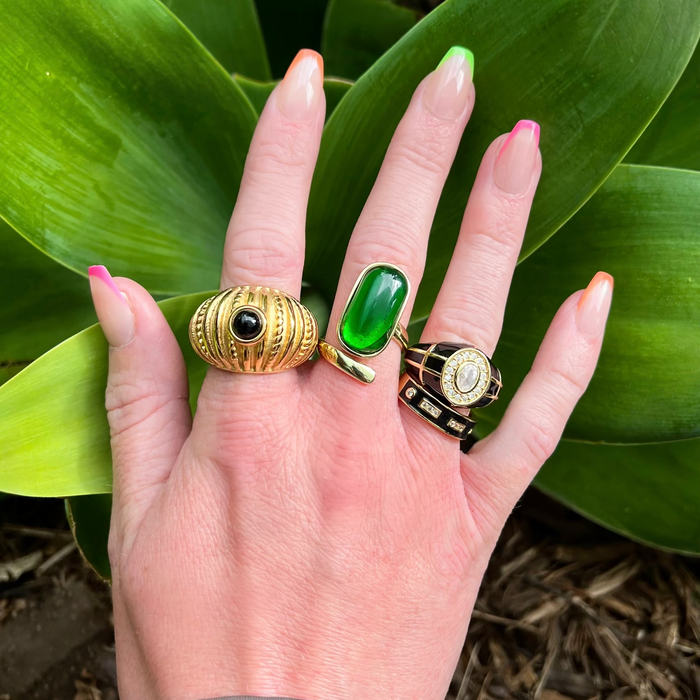 Hand wearing three rings with green gemstones against a leafy background