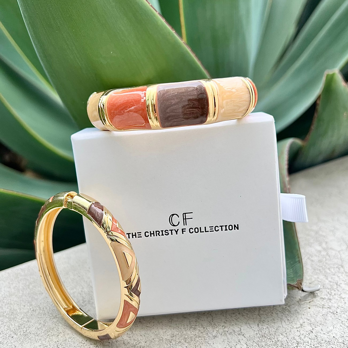 Gold and enamel bangle on a white box with 'The Christy F Collection' branding, against a green plant background.