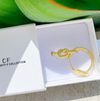 Gold earring in a white box with 'CF' branding, against a green leafy background