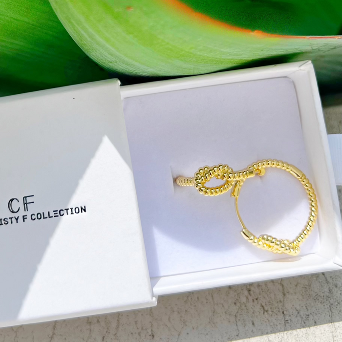 Gold earring in a white box with 'CF' branding, against a green leafy background