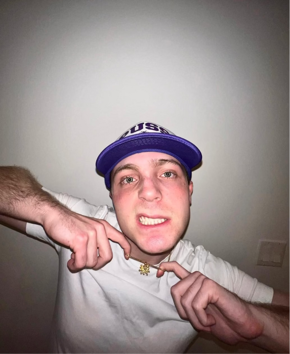 Person wearing a purple cap pointing at themselves against a plain background
