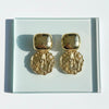 Gold earrings on a clear stand with a light gray background