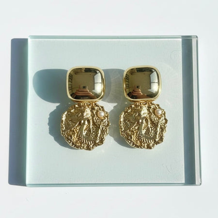Gold earrings on a clear stand with a light gray background