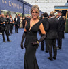 A woman wearing a floor-length black gown with a plunging neckline, long gloves, and a gold necklace stands on a blue carpet at an Emmy event, with photographers and guests in the background.