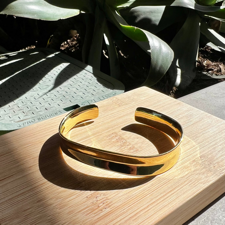 A gold-colored bangle bracelet with a polished finish, displayed on a wooden surface, with a plant in the background.