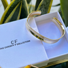 Gold bracelet on a box with 'The Christy F Collection' branding, surrounded by green plants.