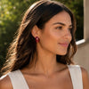 A woman wearing a small, dark red crescent-shaped earring, shown in a close-up profile against a soft-focus outdoor background of green foliage and a light-colored wall.