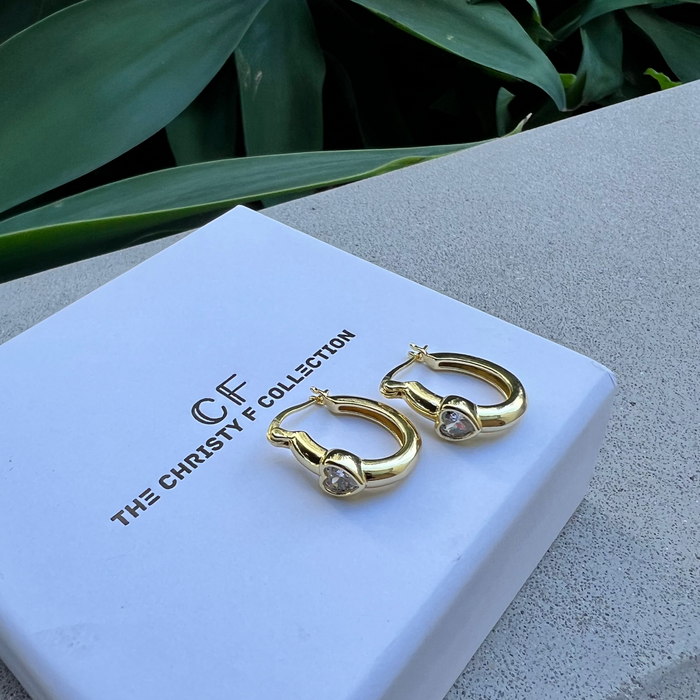 Gold hoop earrings on a white box with 'CF' branding, against a green leafy background.