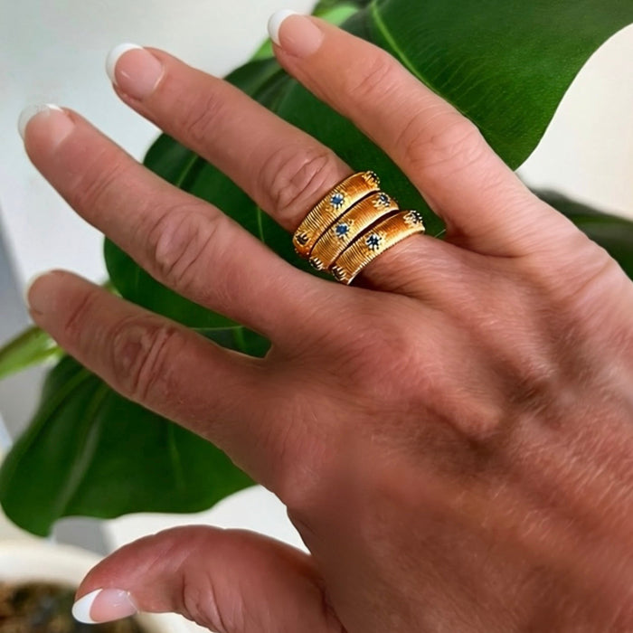 Three gold rings with small blue gemstone accents worn on a person's hand, positioned in front of a large green leaf.