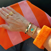 A close-up of a person's hand and wrist wearing multiple gold-toned jewelry pieces, including rings, bracelets, and a turquoise stone pendant, resting on an orange and pink striped fabric surface.