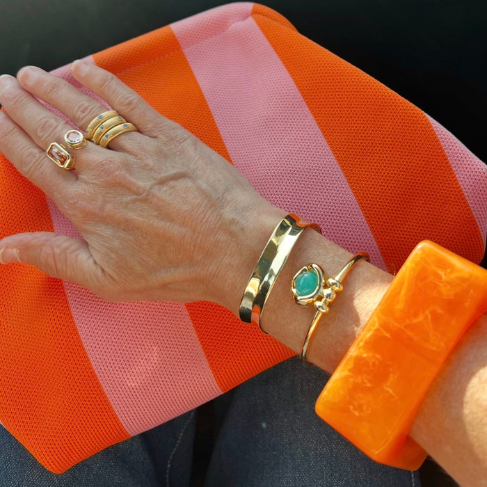 A close-up of a person's hand and wrist wearing multiple gold-toned jewelry pieces, including rings, bracelets, and a turquoise stone pendant, resting on an orange and pink striped fabric surface.