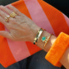 A close-up of a person's hand and wrist wearing multiple gold bracelets, including a wide gold bangle, a slim gold bangle with a turquoise stone, and stacked gold rings, against a bright orange and pink striped fabric background.