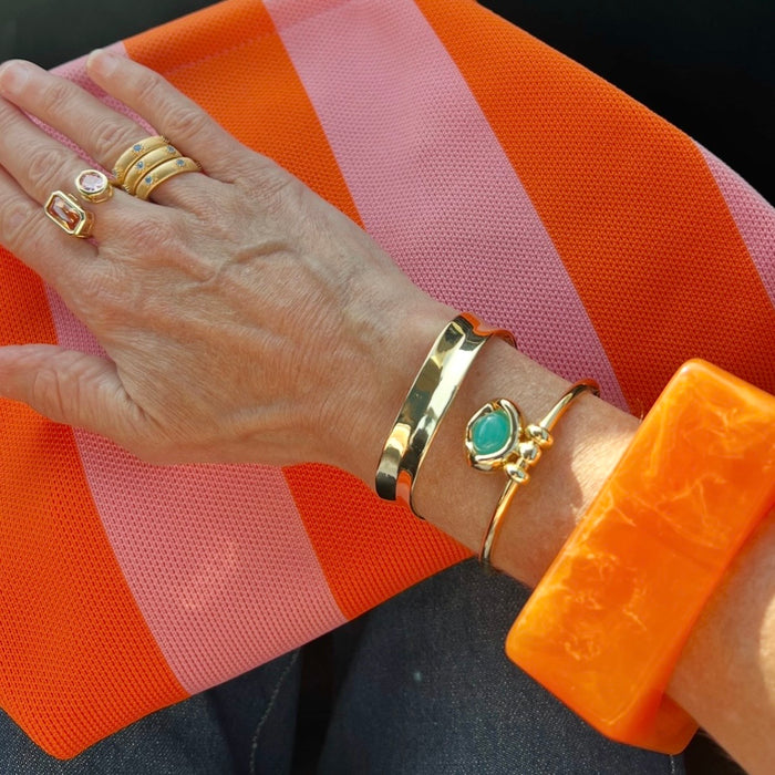 A close-up of a person's hand and wrist wearing multiple gold bracelets, including a wide gold bangle, a slim gold bangle with a turquoise stone, and stacked gold rings, against a bright orange and pink striped fabric background.