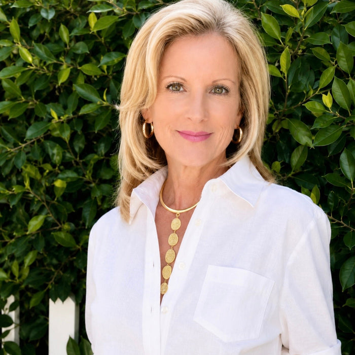 A blonde woman wearing a white button-down shirt and gold jewelry, standing in front of a green leafy hedge.