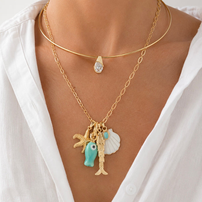 A close-up of a model wearing a layered gold necklace set. The top layer is a delicate gold chain with a small teardrop crystal pendant.