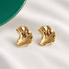 A pair of gold-toned, organic-shaped stud earrings with a textured, wavy surface, displayed on a white fabric background with a green plate edge visible in the top left corner.