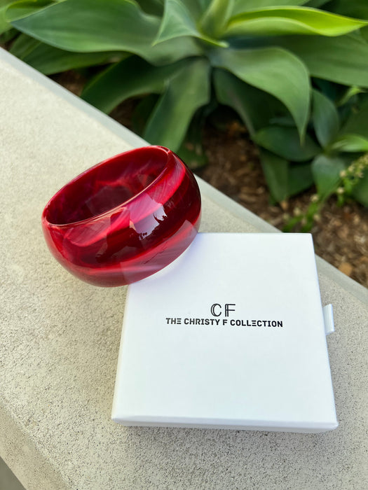 Red bracelet and white box with 'CF The Christy F Collection' branding on a textured surface with greenery in the background.