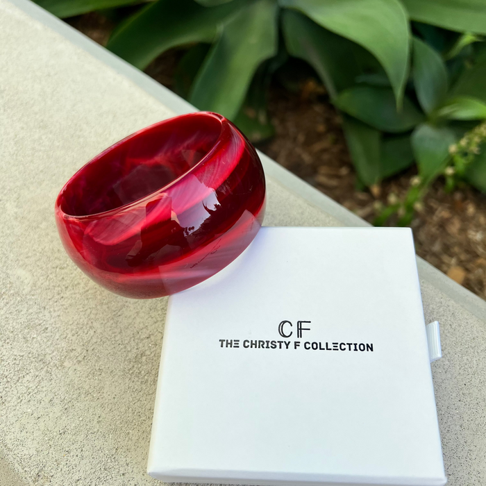 Red bracelet and white box with 'CF The Christy F Collection' branding on a textured surface with greenery in the background.