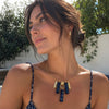 A woman wearing a gold necklace with a rectangular pendant featuring blue and white patterned lapis stones, photographed outdoors against a white wall and green foliage.