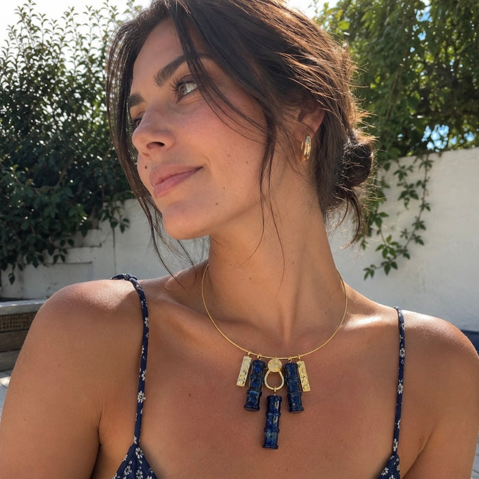A woman wearing a gold necklace with a rectangular pendant featuring blue and white patterned lapis stones, photographed outdoors against a white wall and green foliage.