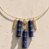 A gold-tone necklace featuring five hanging charms: two blue lapis lazuli cylinder pendants, two textured gold bar charms with small clear stones, and one round gold medallion, displayed on a textured beige stone surface.