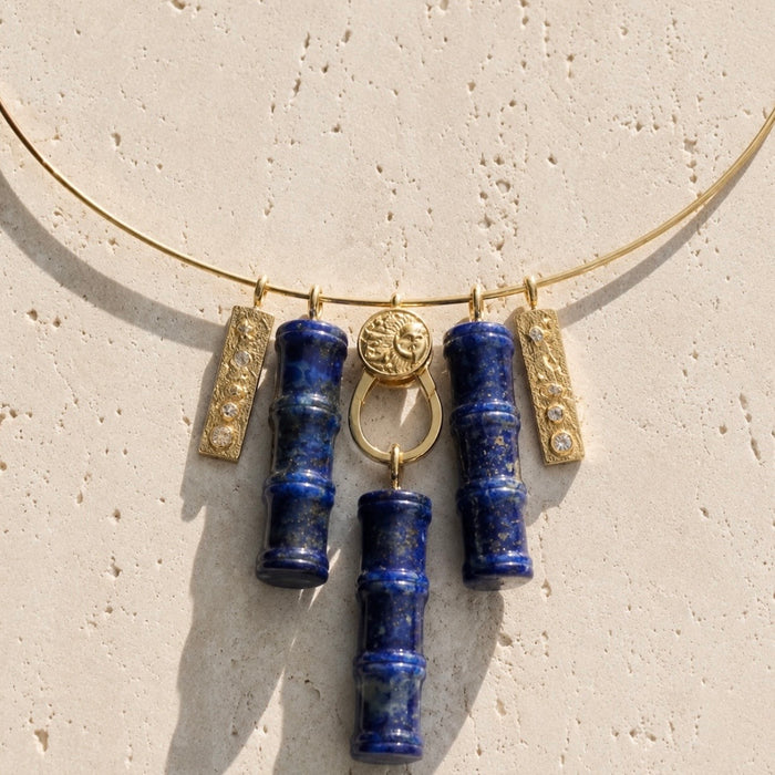 A gold-tone necklace featuring five hanging charms: two blue lapis lazuli cylinder pendants, two textured gold bar charms with small clear stones, and one round gold medallion, displayed on a textured beige stone surface.