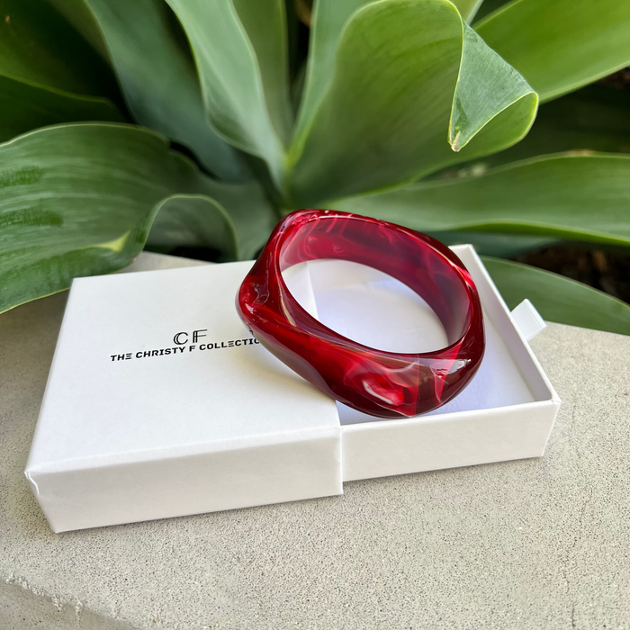 Red bracelet in a white box with 'CF' branding, set against a green leafy background.