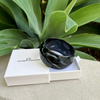 Black bracelet in a white box with 'CF Collection' branding, placed on a stone surface with green foliage in the background.