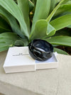 Black bracelet in a white box with 'CF Collection' branding, placed on a stone surface with green foliage in the background.