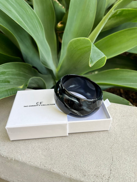 Black bracelet in a white box with 'CF Collection' branding, placed on a stone surface with green foliage in the background.