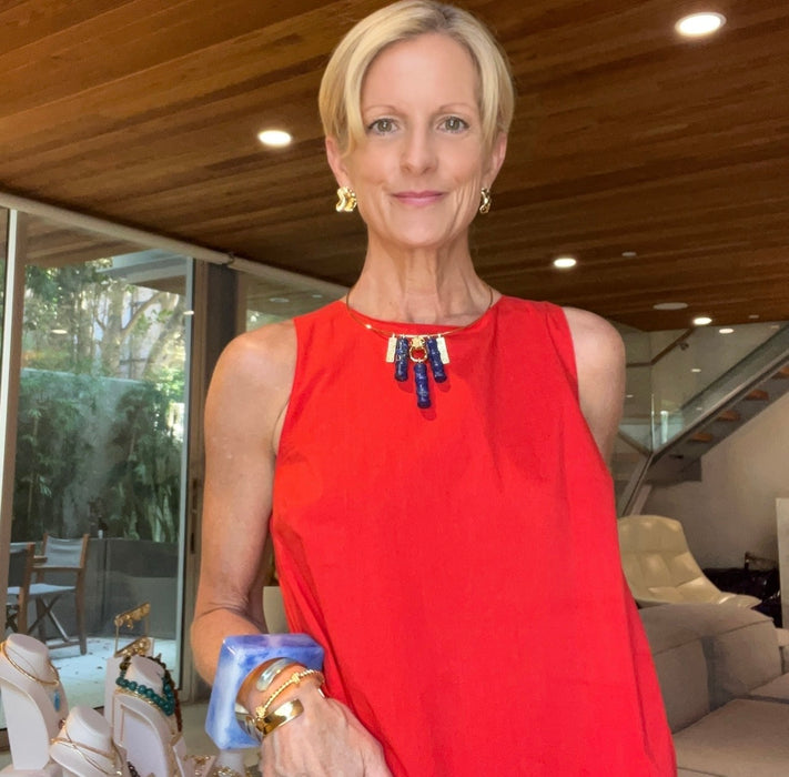 A woman with short blonde hair wearing a bright red sleeveless top and gold jewelry, holding a blue and white box in a modern indoor setting with a wooden ceiling.