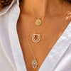 A close-up of a person wearing layered gold necklaces with distinct pendants: a round coin-style pendant, a horseshoe-shaped pendant set with clear stones, and a Hamsa hand pendant also accented with clear stones, styled with a white shirt.