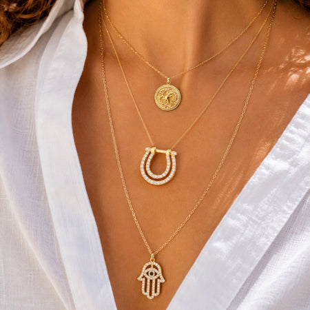 A close-up of a person wearing layered gold necklaces with distinct pendants: a round coin-style pendant, a horseshoe-shaped pendant set with clear stones, and a Hamsa hand pendant also accented with clear stones, styled with a white shirt.