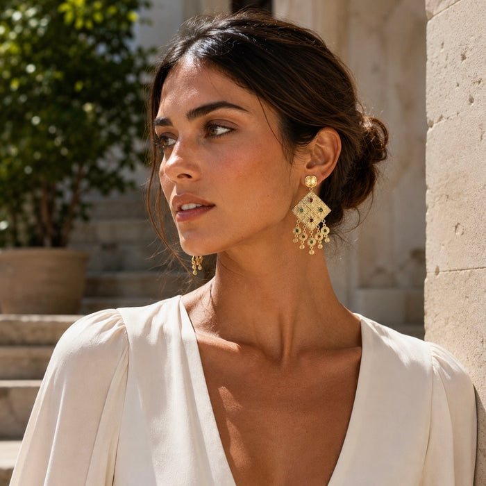 A woman wearing ornate gold dangle earrings with geometric filigree and hanging charms, styled with a cream-colored blouse in an outdoor setting.