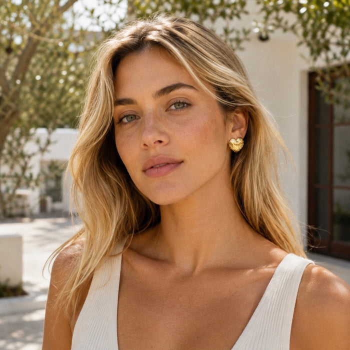 A blonde woman wearing a small gold heart-shaped stud earring, photographed outdoors in bright natural light with trees and a white building in the background.