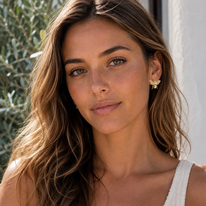 A close-up portrait of a woman with long wavy brown hair wearing a small gold bee stud earring, set against a soft-focus background of green foliage and a white wall.