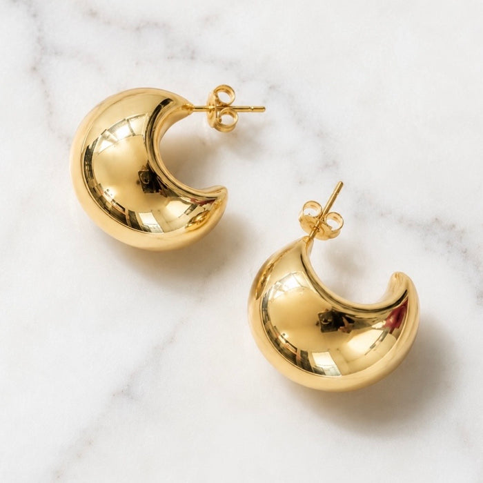 A pair of polished gold crescent moon stud earrings resting on a white marble surface.