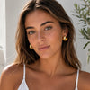 A close-up of a woman with brown hair wearing gold crescent-shaped hoop earrings and a white spaghetti-strap top, set against a bright, sunlit neutral background with a leafy plant on the right.