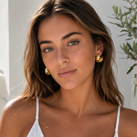 A close-up of a woman with brown hair wearing gold crescent-shaped hoop earrings and a white spaghetti-strap top, set against a bright, sunlit neutral background with a leafy plant on the right.