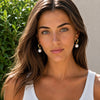 A woman with long brown hair wearing gold hoop earrings with white pearl drops and a delicate gold chain necklace, photographed outdoors against green foliage and a light wall.